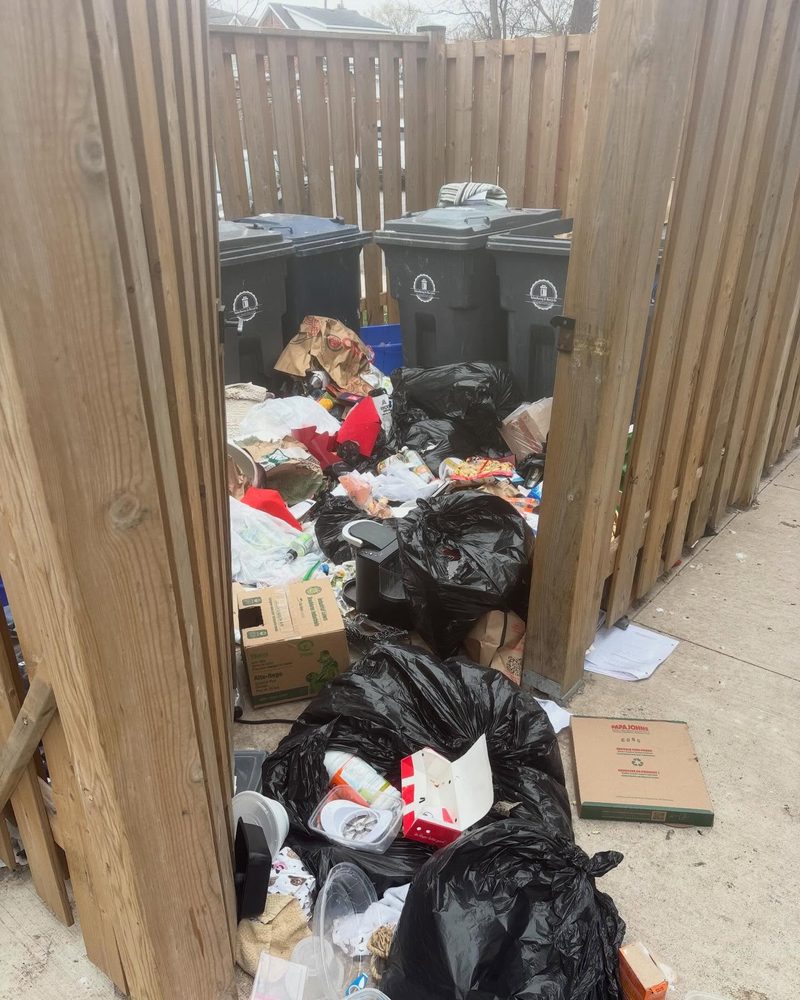 A fenced bin enclosure overflowing with loose garbage bags and boxes around the collection bins