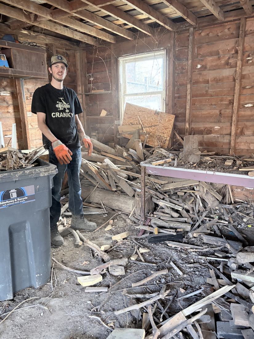 A crew member standing inside an old shed filled knee-deep with broken lumber and scrap wood