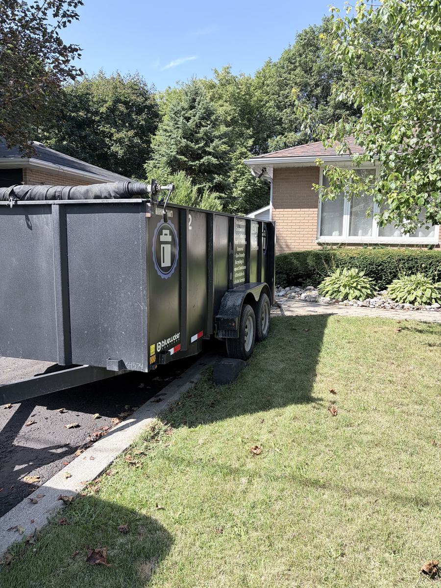 A bin backed up along a driveway beside a brick bungalow on a clear summer day