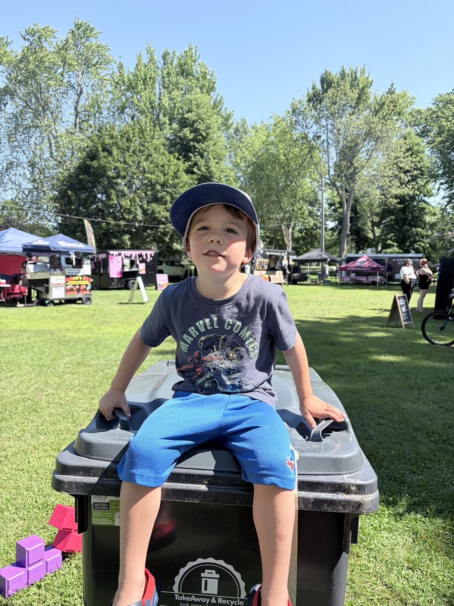 A child sitting on top of a branded TakeAway & Recycle bin at a summer community event
