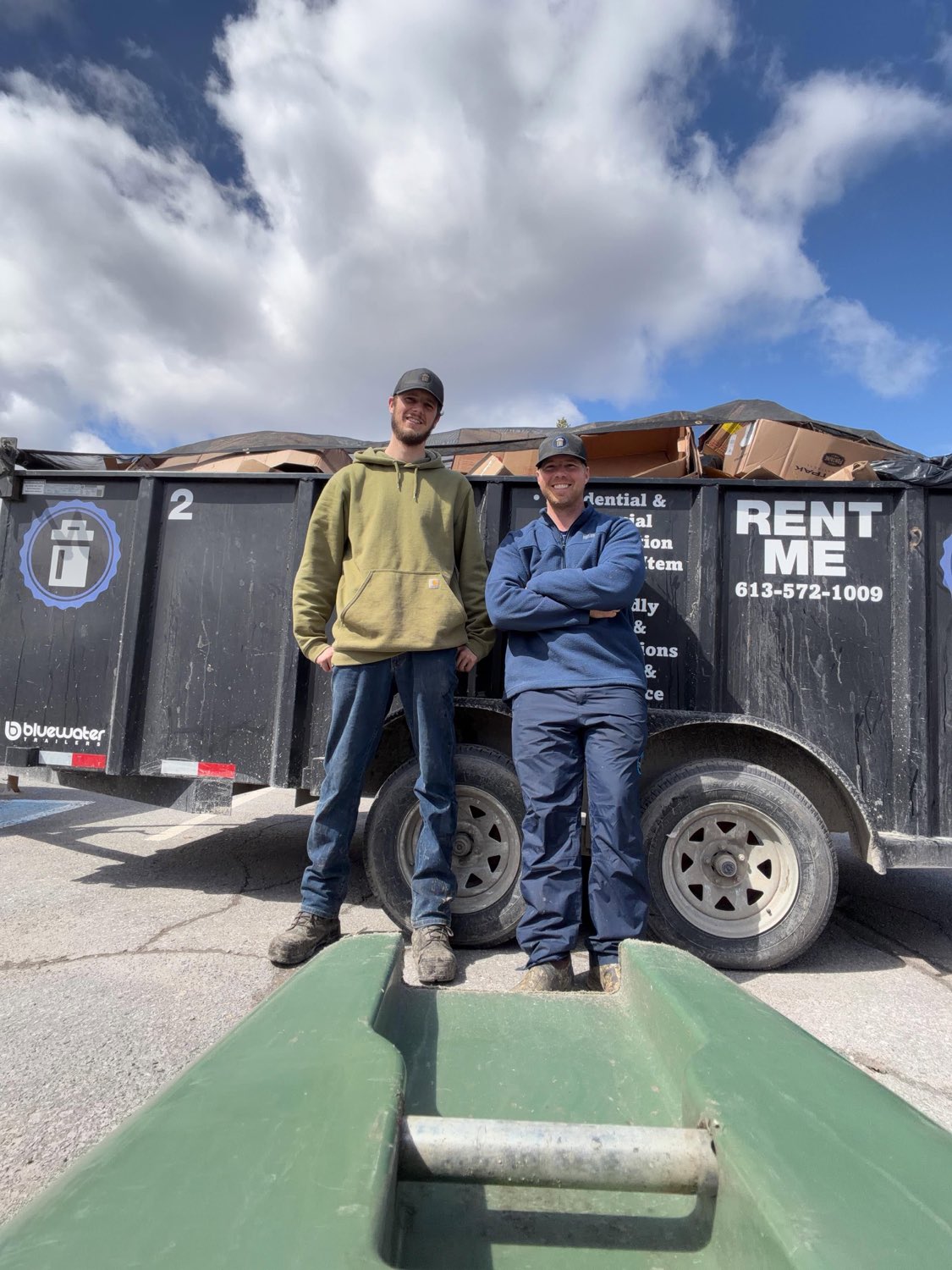 Two crew members standing in front of a loaded Rent Me dump trailer full of flattened cardboard