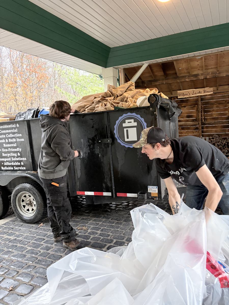 Two crew members loading bags and a rolled tarp into the black dump trailer under the covered porch of a smokehouse restaurant