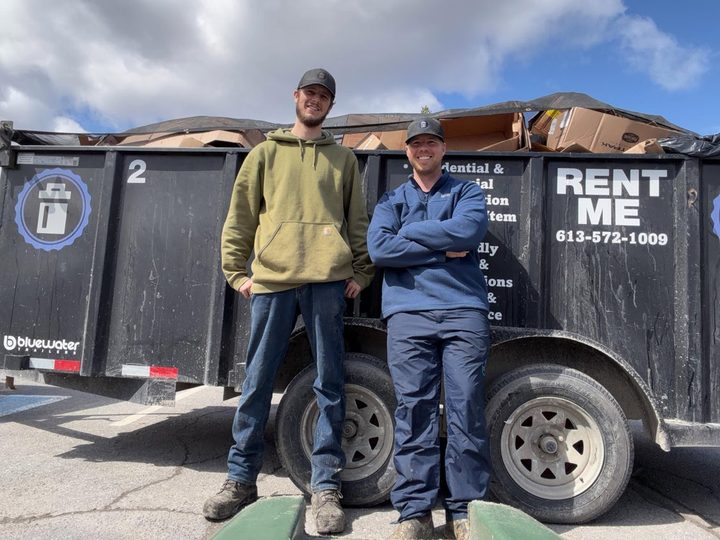 Two crew members standing in front of the black 18-yard RENT ME dump trailer loaded with cardboard