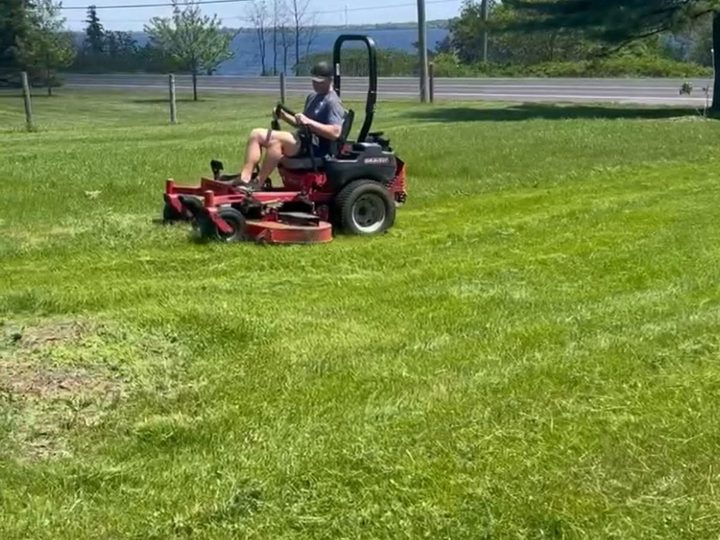 A crew member on a zero-turn mower cutting a large open lawn on a bright day outside Kingston